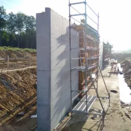 Wysoka, szara ściana oporowa z betonu, w trakcie budowy, wsparta rusztowaniem metalowym, widoczne ślady szalunku, w tle ziemia i drzewa.