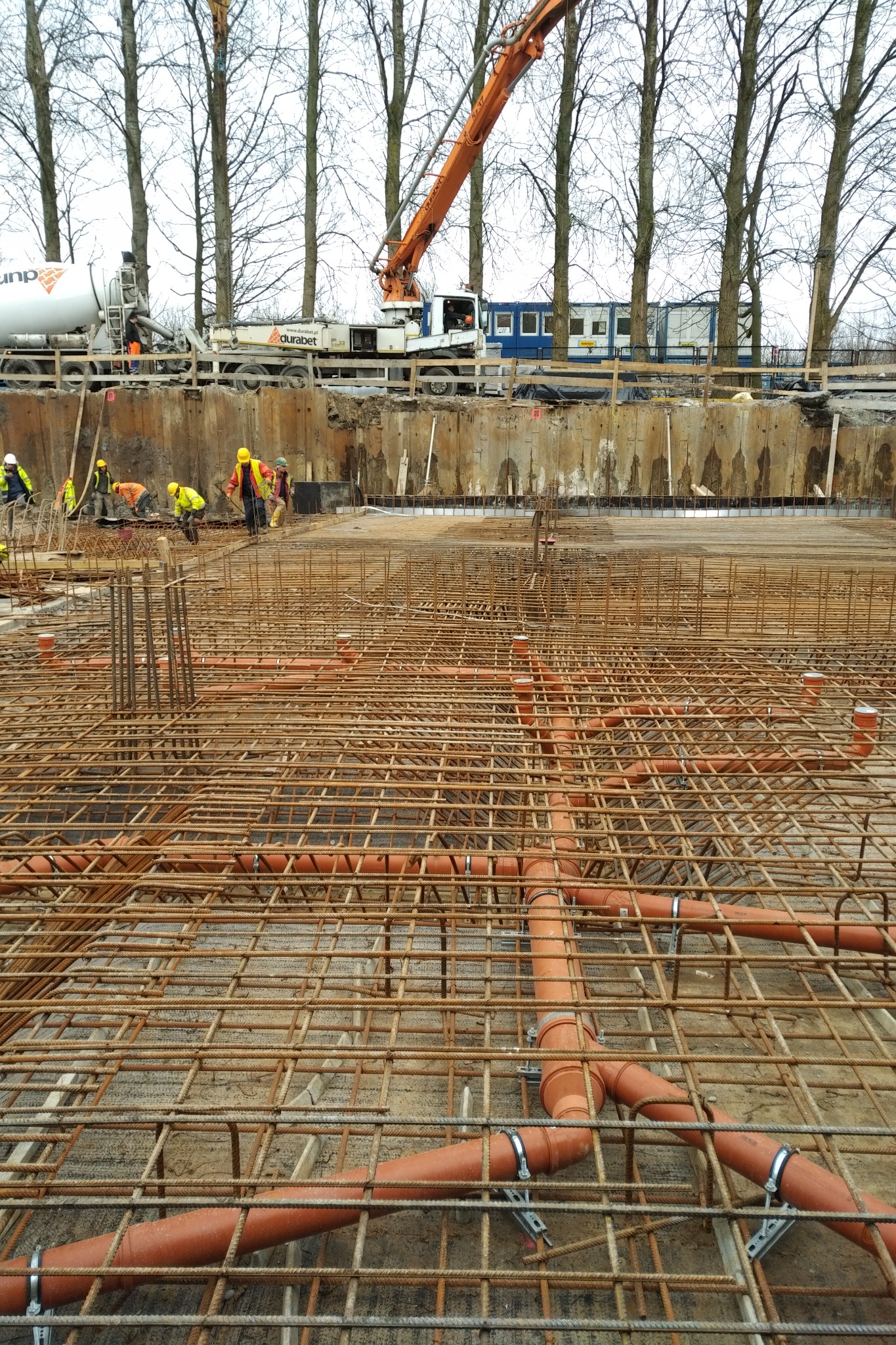 Siatka zbrojeniowa z rurami kanalizacyjnymi w fundamencie, w tle pracownicy budowlani i maszyny na placu budowy.