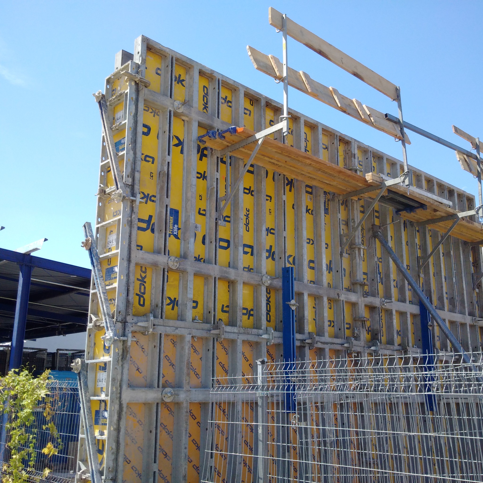 Szare szalunki Doka z żółtymi wkładami na budowie, widoczne rusztowanie i zbrojenia, niebieskie niebo w tle.