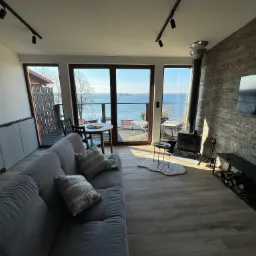 Nowoczesny apartament z widokiem na jezioro, jasne wnętrze z szarą sofą, kamienną ścianą z kominkiem i telewizorem, oraz panoramicznymi oknami.