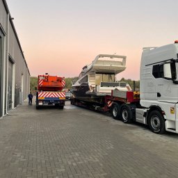 Radlight - Transport jachtu na lawecie, z pomarańczowym pojazdem pilotującym z tyłu. Szary budynek magazynowy w tle, różowe niebo o zmierzchu. Widok z boku, na zewnątrz.