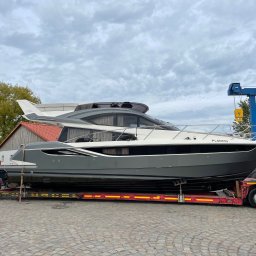 Radlight - Transport jachtu motorowego na niskopodwoziowej lawecie, widoczny fragment suwnicy bramowej i pracownik zabezpieczający ładunek. Szary jacht z białymi elementami.