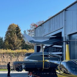 Radlight - Hangar z łodziami i helikopterem, mężczyzna w ciemnym ubraniu stoi obok. Żółty helikopter na pierwszym planie, łodzie w hangarze w tle, błękitne niebo.