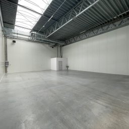 Radlight - Pusta hala magazynowa z betonową posadzką i oświetleniem dachowym w Giżycku. Białe ściany z paneli, widoczne instalacje wentylacyjne pod sufitem.