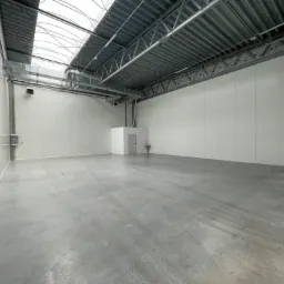 Pusta hala magazynowa z betonową posadzką i oświetleniem dachowym w Giżycku. Białe ściany z paneli, widoczne instalacje wentylacyjne pod sufitem.