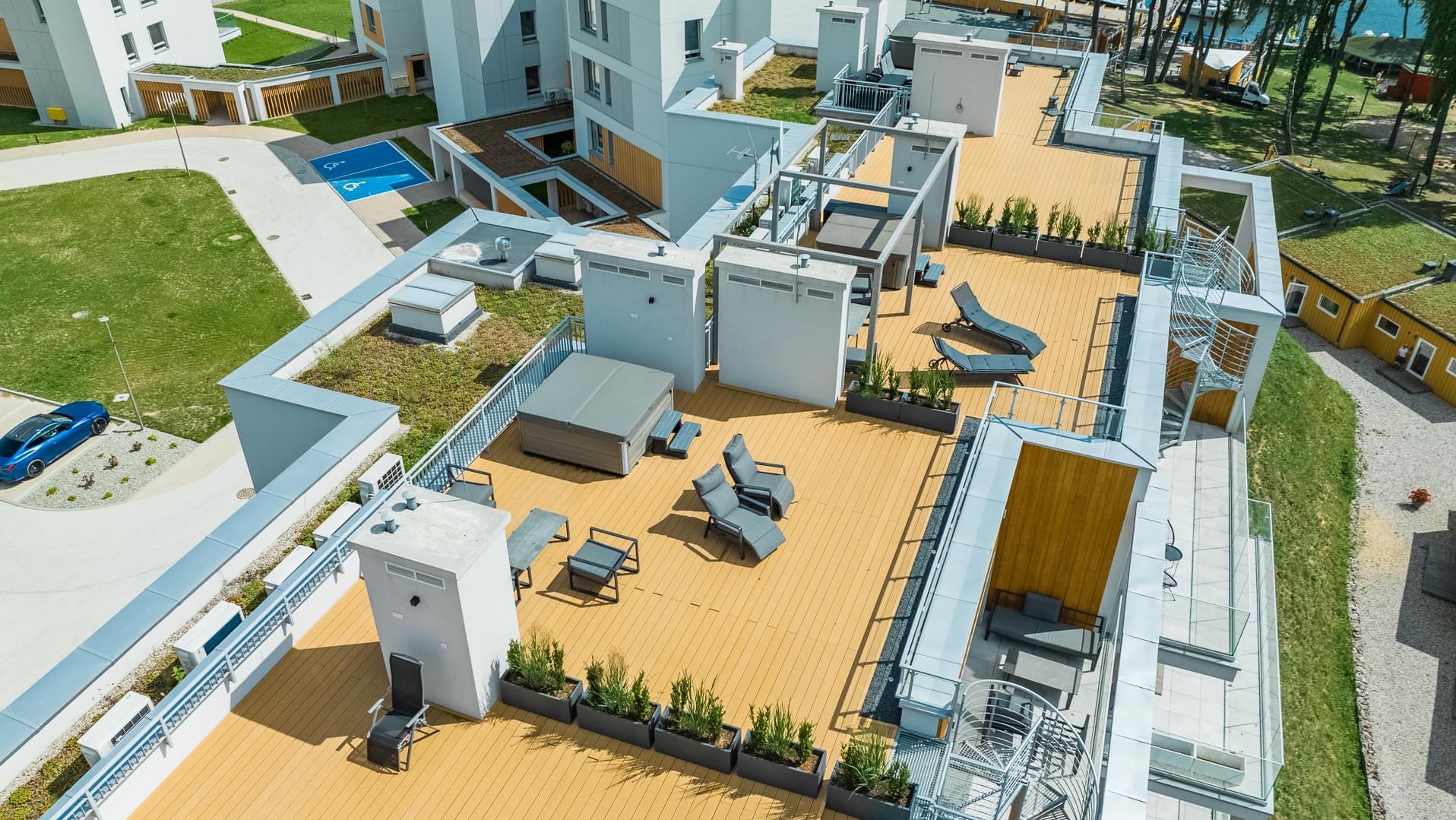 Widok z lotu ptaka na taras apartamentów Stranda Residence z jacuzzi, leżakami, meblami wypoczynkowymi i spiralnymi schodami, otoczony zielenią i budynkami o nowoczesnej architekturze.