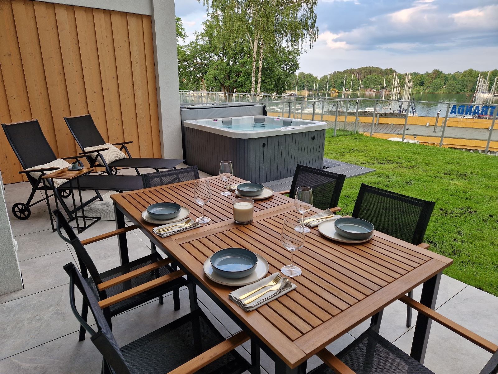 Zewnętrzny taras z drewnianym stołem nakrytym dla czterech osób, jacuzzi i leżakami w tle, widok na jezioro z pomostem i zacumowanymi jachtami.