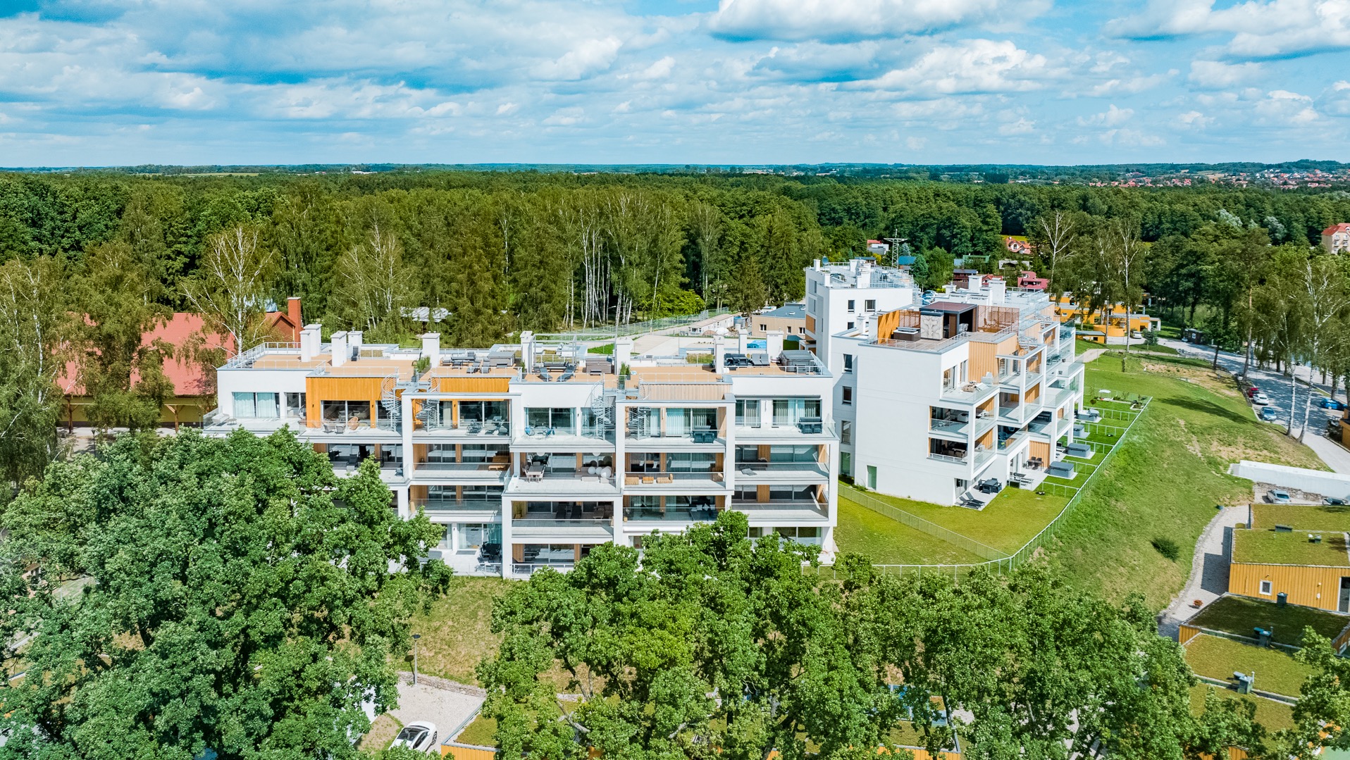 Wielopiętrowy apartamentowiec z balkonami, otoczony zielenią drzew i trawników, widok z lotu ptaka.