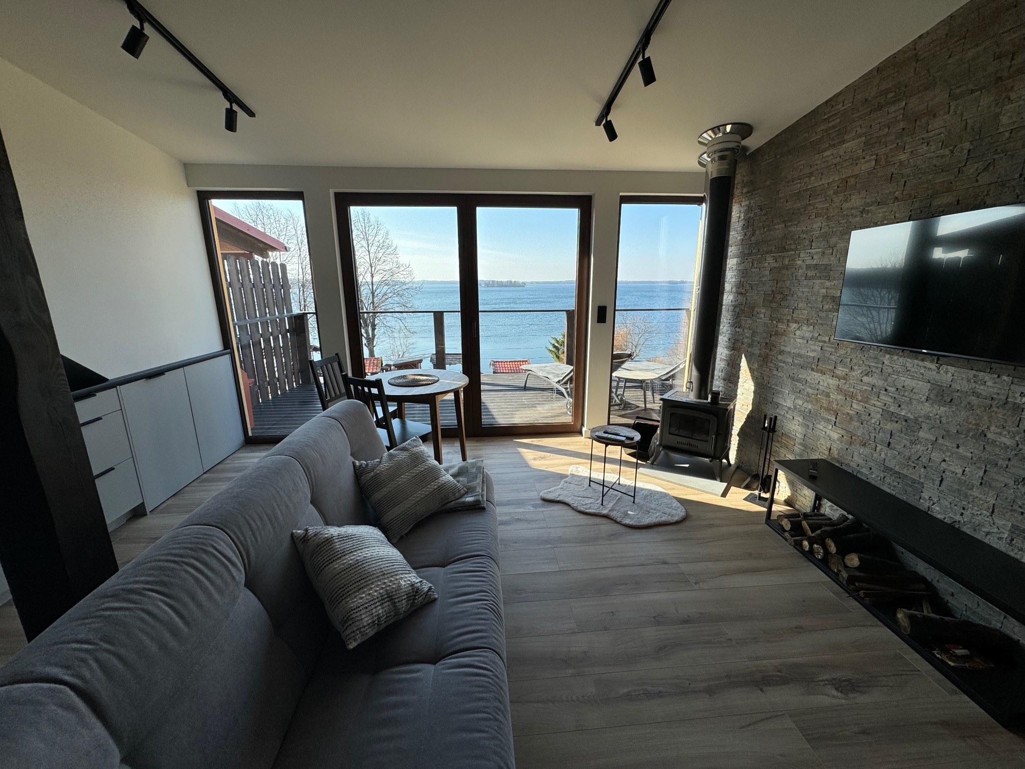 Nowoczesny apartament z widokiem na jezioro, jasne wnętrze z szarą sofą, kamienną ścianą z kominkiem i telewizorem, oraz panoramicznymi oknami.