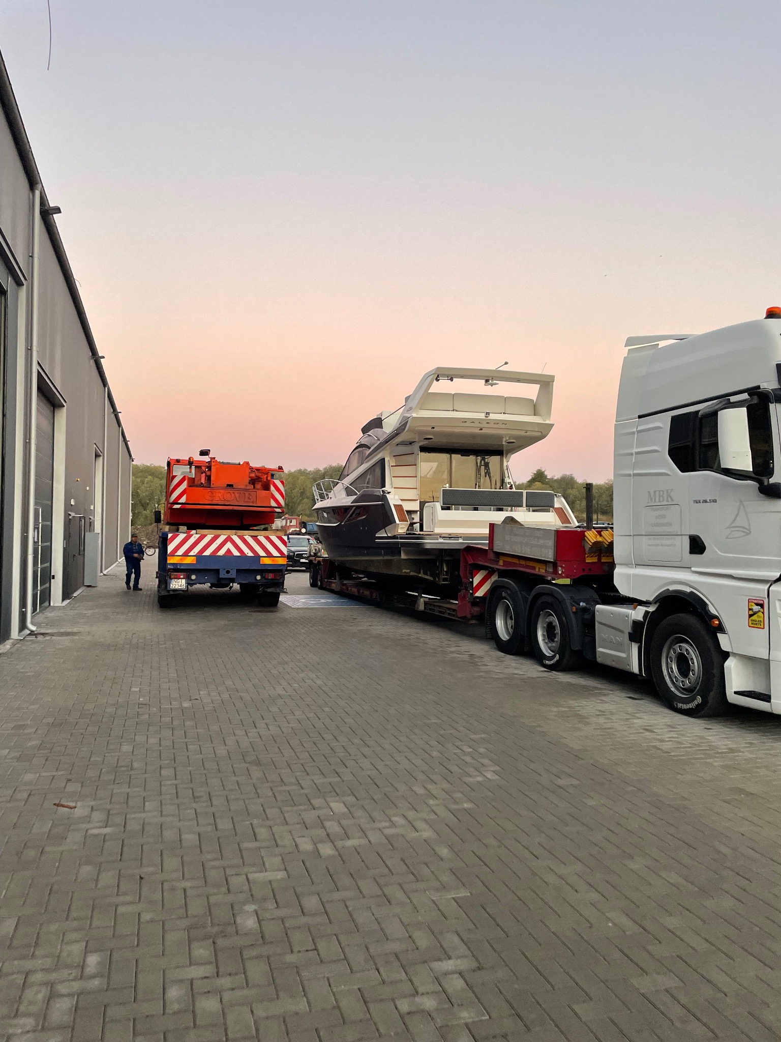 Transport jachtu na lawecie, z pomarańczowym pojazdem pilotującym z tyłu. Szary budynek magazynowy w tle, różowe niebo o zmierzchu. Widok z boku, na zewnątrz.