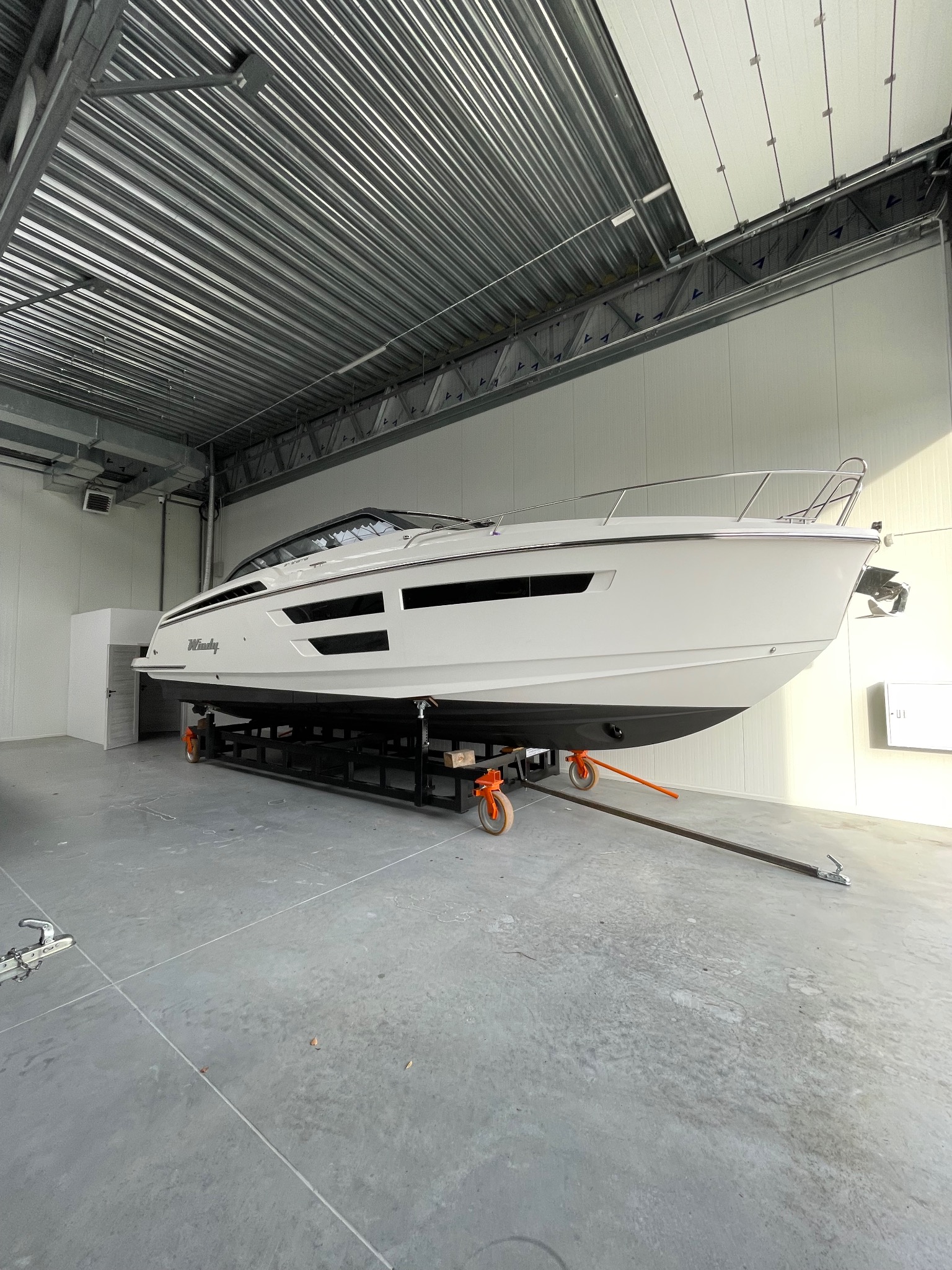 Biała motorówka Windy na wózku transportowym w hali z blachy falistej. Jacht gotowy do sezonu, zabezpieczony w magazynie.