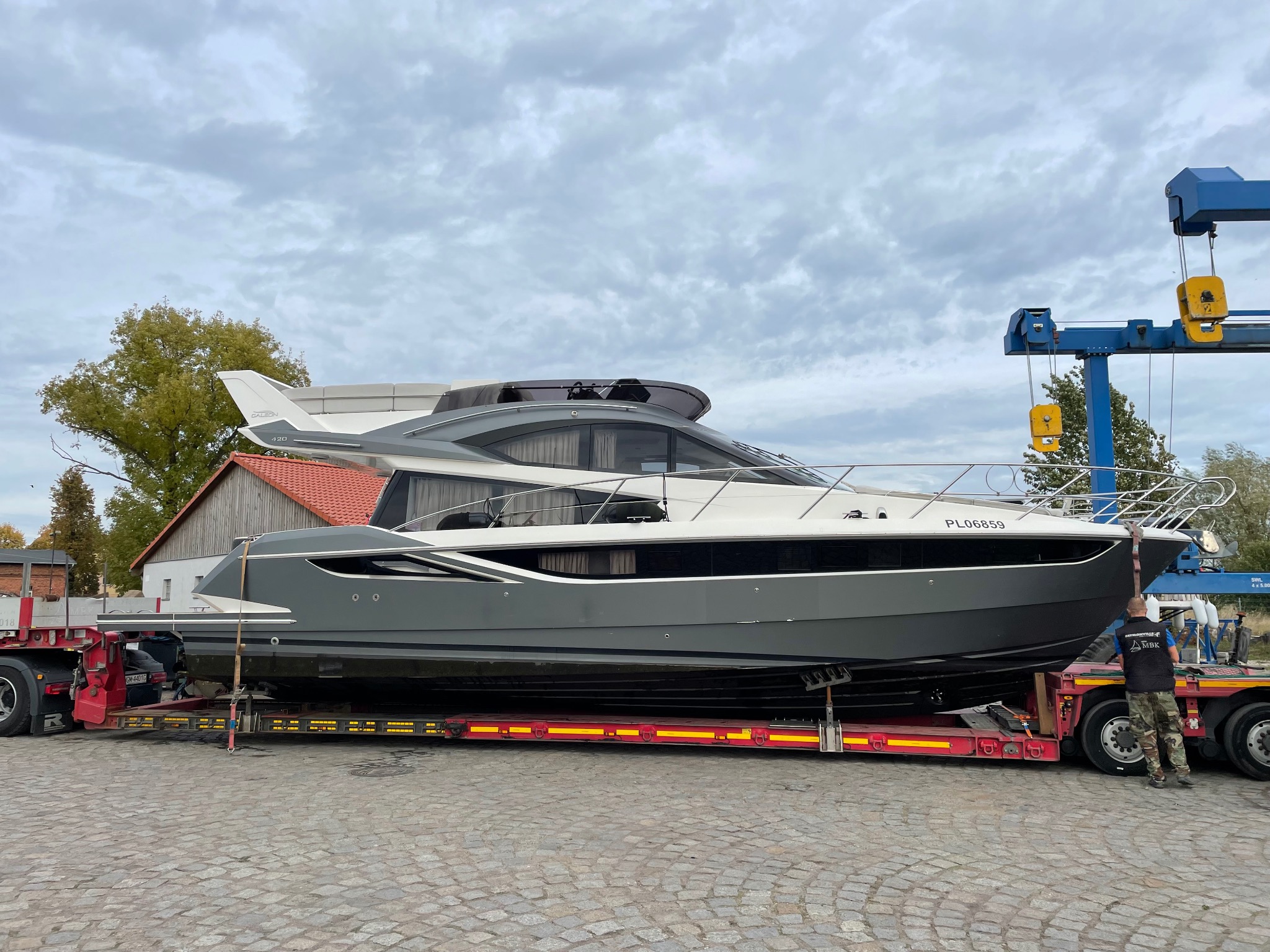 Transport jachtu motorowego na niskopodwoziowej lawecie, widoczny fragment suwnicy bramowej i pracownik zabezpieczający ładunek. Szary jacht z białymi elementami.