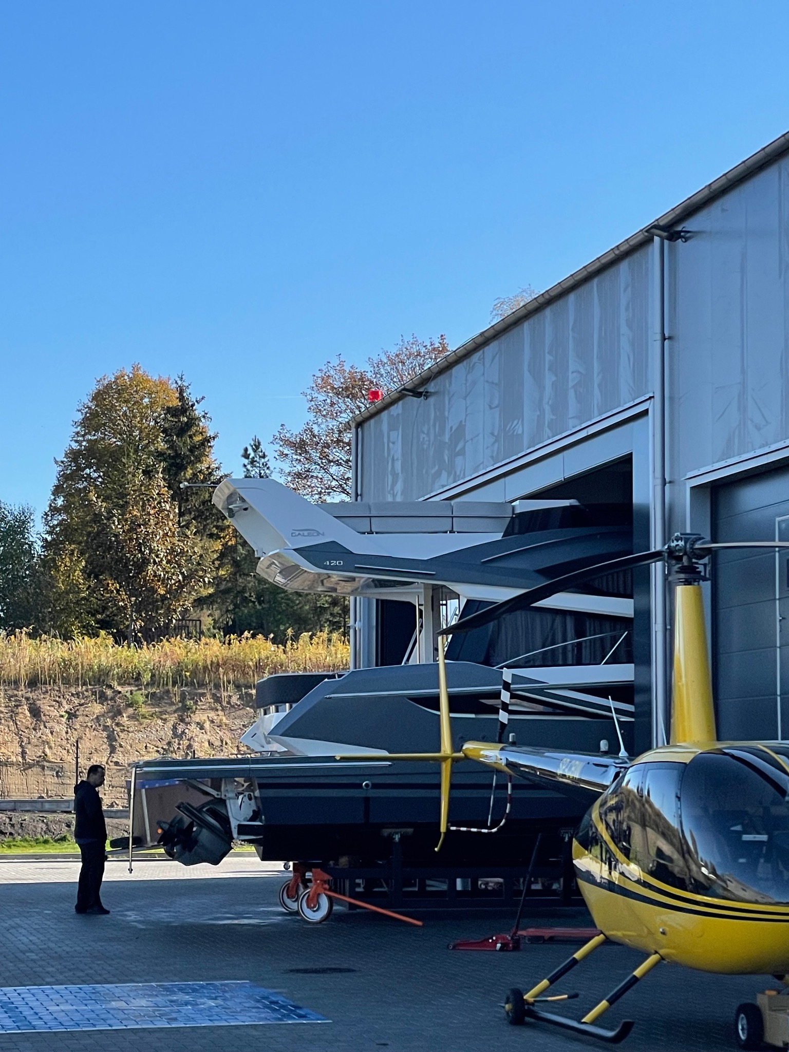 Hangar z łodziami i helikopterem, mężczyzna w ciemnym ubraniu stoi obok. Żółty helikopter na pierwszym planie, łodzie w hangarze w tle, błękitne niebo.