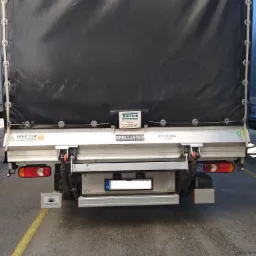 Tył samochodu dostawczego z czarną plandeką, widoczne logo firm Wektor, Turplan i Dhollandia, tablica rejestracyjna z Polską flagą, oraz żółte pasy na betonowej nawierzchni.