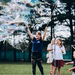 Uśmiechnięta kobieta w koszulce z napisem 'Animator' tworzy ogromne bańki mydlane na imprezie plenerowej, dzieci obserwują z zaciekawieniem.