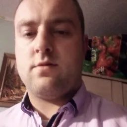 Portret mężczyzny w fioletowej koszuli, na tle ściany i szafki z produktami spożywczymi.