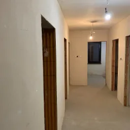 Korytarz w stanie surowym po nałożeniu wstępnych warstw tynku, widoczne ościeżnice z cegły i prowizoryczne oświetlenie.