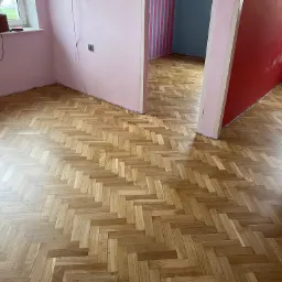 Parkiet ułożony w jodełkę w przestronnym, odświeżanym wnętrzu z różowymi i czerwonymi ścianami, widoczne ślady po malowaniu i narzędzia na parapecie.