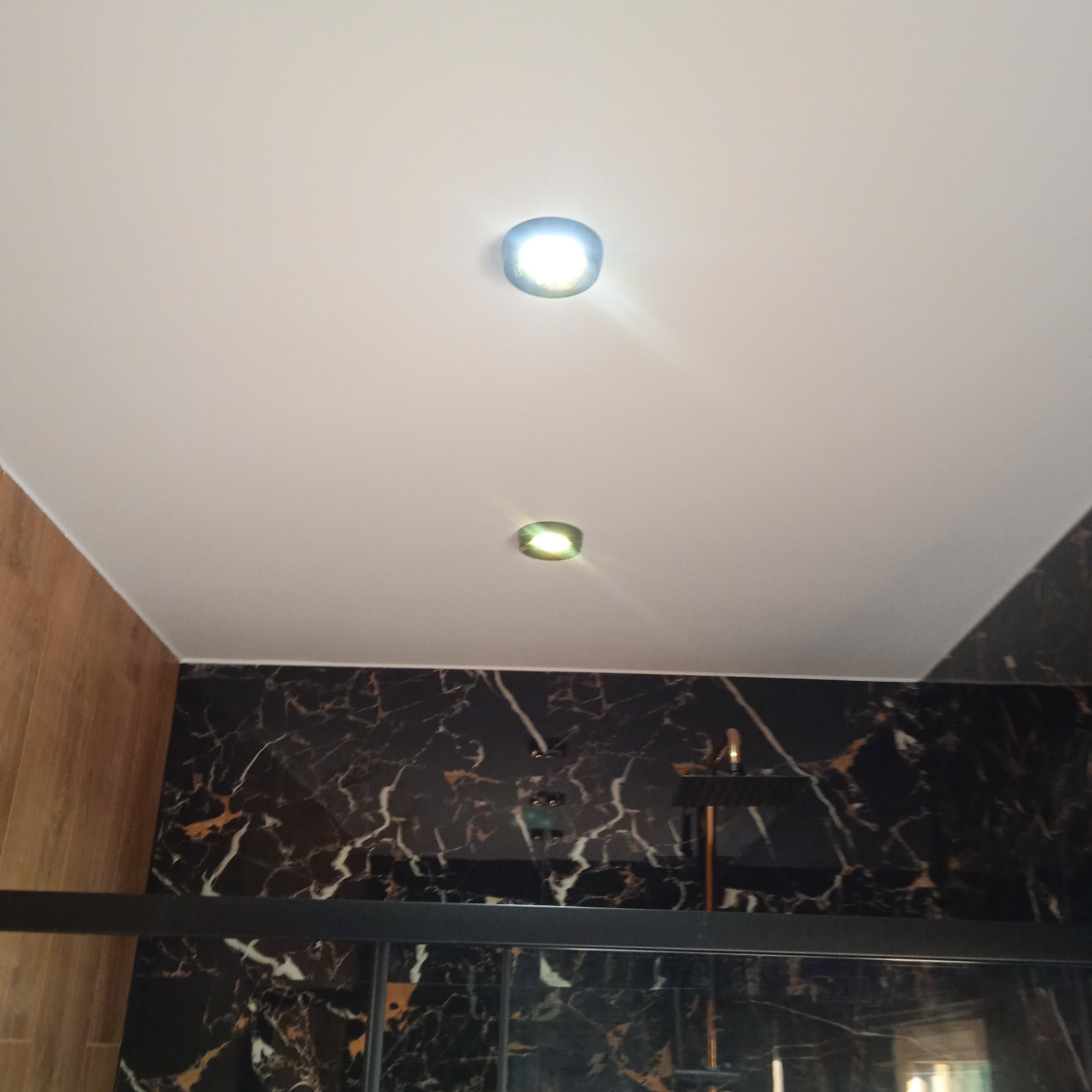 Wnętrze łazienki z czarną marmurową ścianą, widoczne dwa punktowe źródła światła w białym suficie i fragment kabiny prysznicowej z czarną ramą.