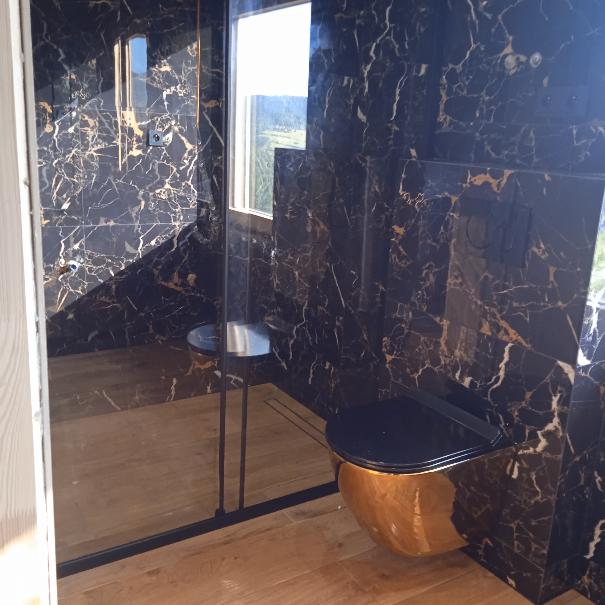 Luksusowa łazienka z czarnymi marmurowymi płytkami ze złotymi żyłami, złotą wiszącą muszlą klozetową i drewnianą podłogą, widok na okno z krajobrazem.