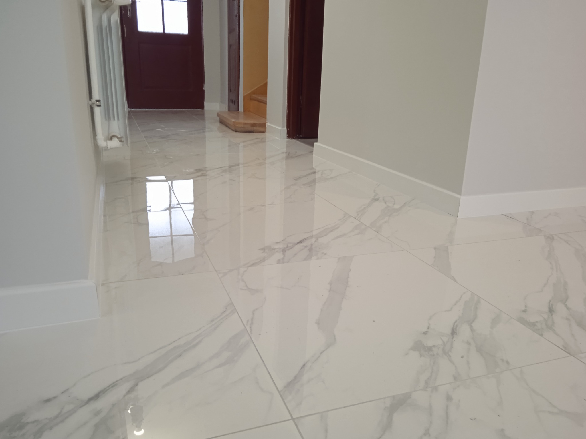 Błyszcząca, biała marmurowa podłoga w korytarzu z widocznym grzejnikiem i drzwiami po lewej stronie oraz fragmentem schodów w tle.