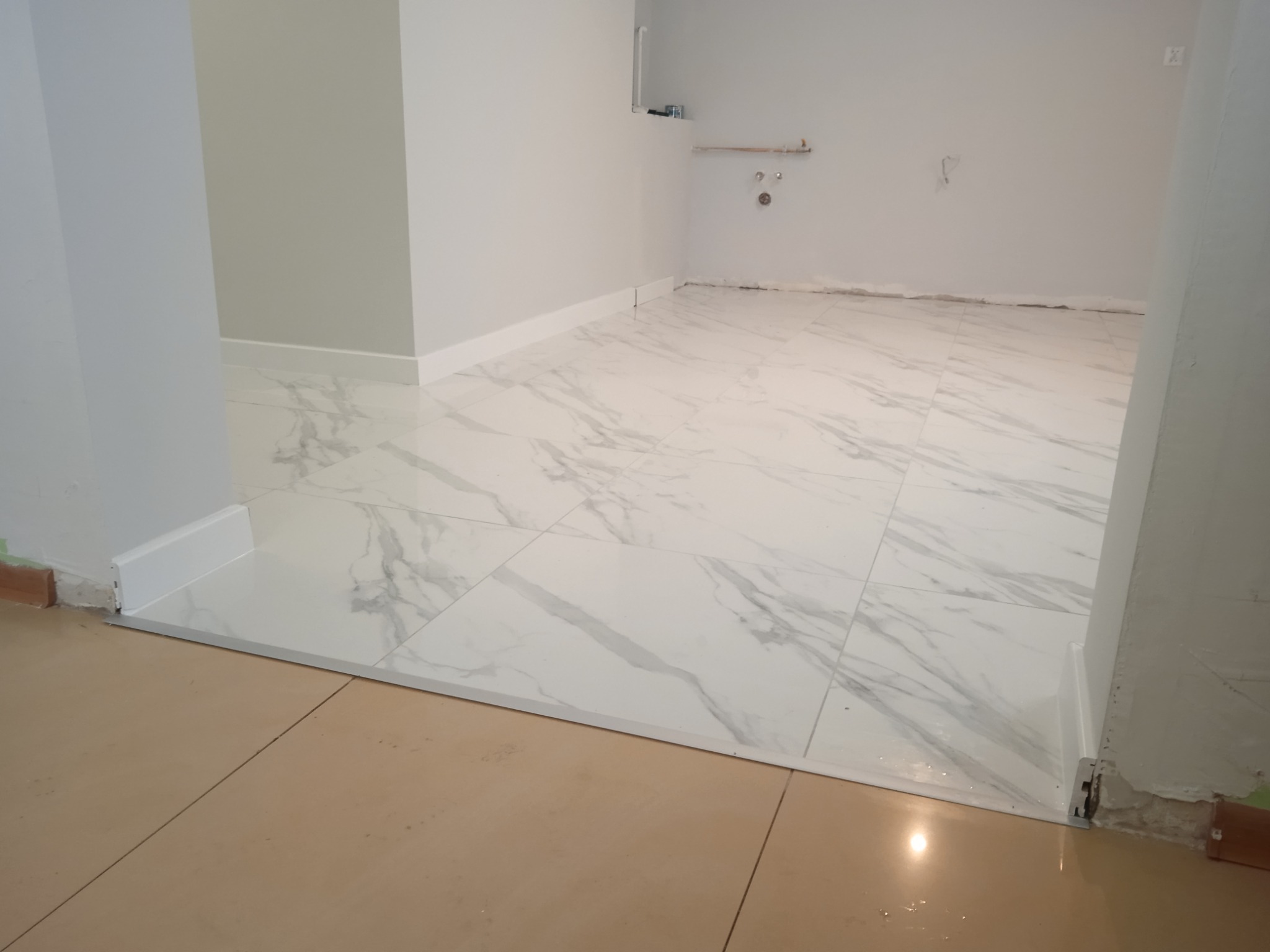 Wykończone wnętrze z białymi płytkami podłogowymi imitującymi marmur, białe listwy przypodłogowe i widoczne niedoskonałości wykończenia przy ścianach.