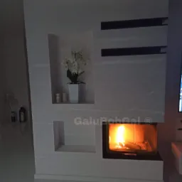 Nowoczesny kominek w zabudowie z białego kamienia z wnękami dekoracyjnymi, w jednej z nich biała orchidea w doniczce, widoczne płomienie.