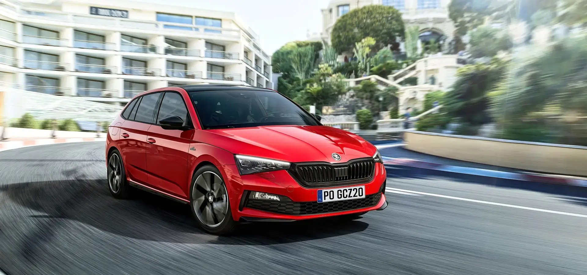 Dynamiczne ujęcie czerwonej Skody Scala z czarnym dachem, pędzącej po krętej drodze z rozmytym tłem luksusowej zabudowy i zieleni.