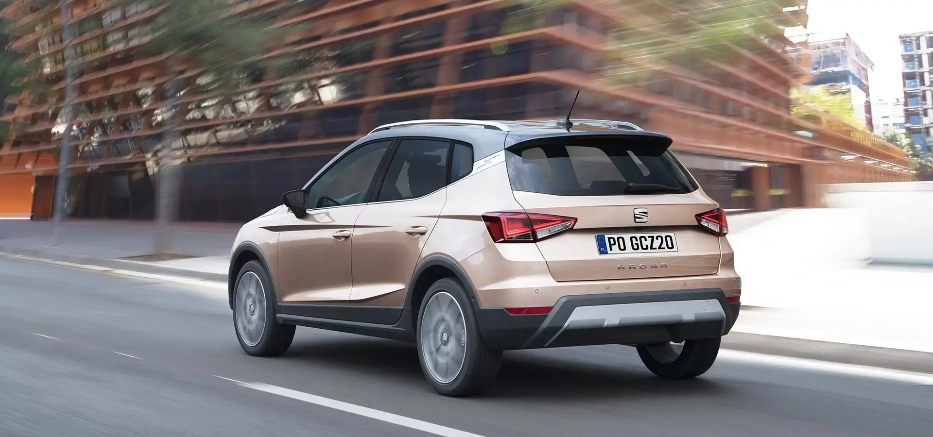 Złoty Seat Arona, widok z tyłu, w ruchu na tle nowoczesnego budynku biurowego z poziomymi pasami. Tablica rejestracyjna PO GCZ20.