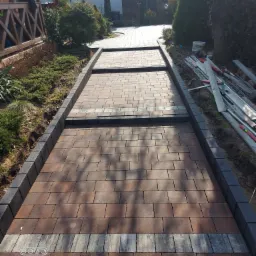 Ułożona ścieżka z kostki brukowej w odcieniach brązu i szarości, obrzeża z ciemnej kostki, widok z perspektywy pieszego, trawnik po bokach, w tle dom i drzewa.