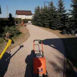 Pomarańczowa zagęszczarka płytowa na żwirowej ścieżce w ogrodzie, w tle dom i pracownicy podczas robót ziemnych.