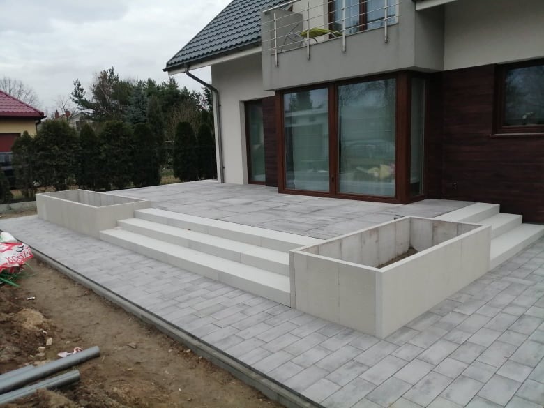 Szara kostka brukowa ułożona na tarasie z donicami i schodami, widok na dom z brązowymi oknami.