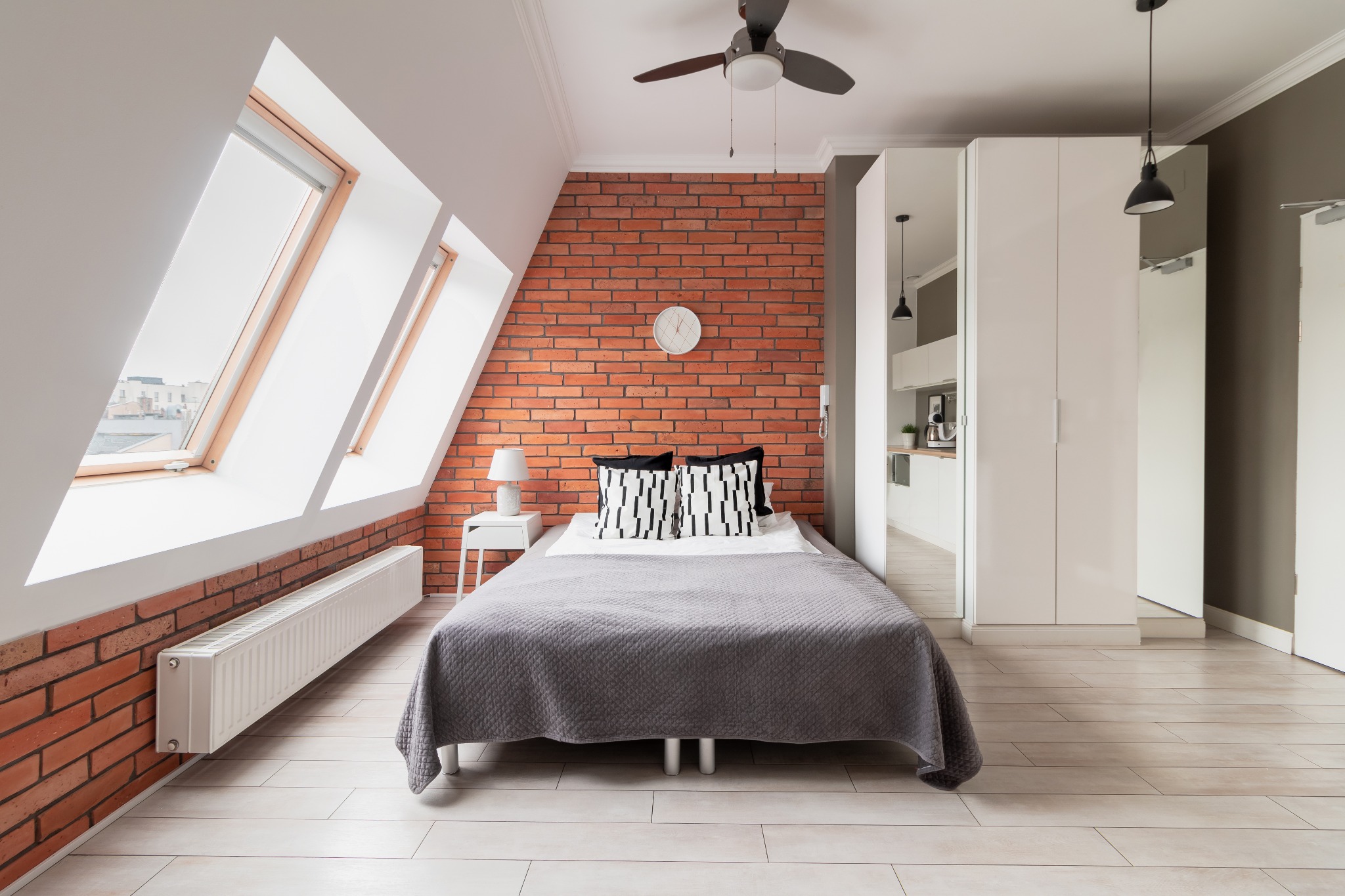 Jasna sypialnia na poddaszu z ceglaną ścianą, oknami dachowymi, szarą narzutą na łóżku i wentylatorem sufitowym.