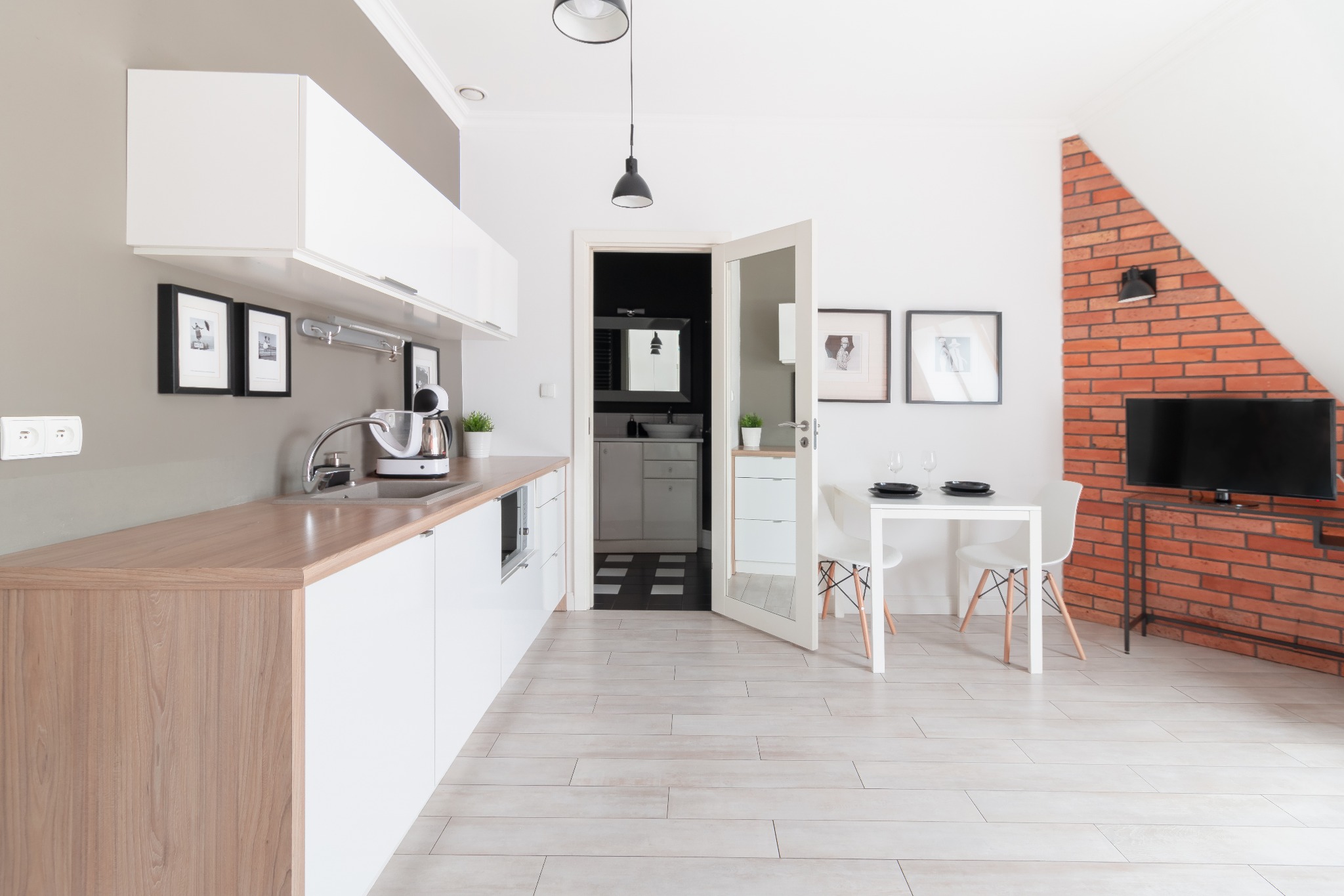 Jasne wnętrze aneksu kuchennego z białymi szafkami, drewnianym blatem, stołem z krzesłami i ceglaną ścianą z telewizorem w tle, widok na łazienkę przez otwarte drzwi.