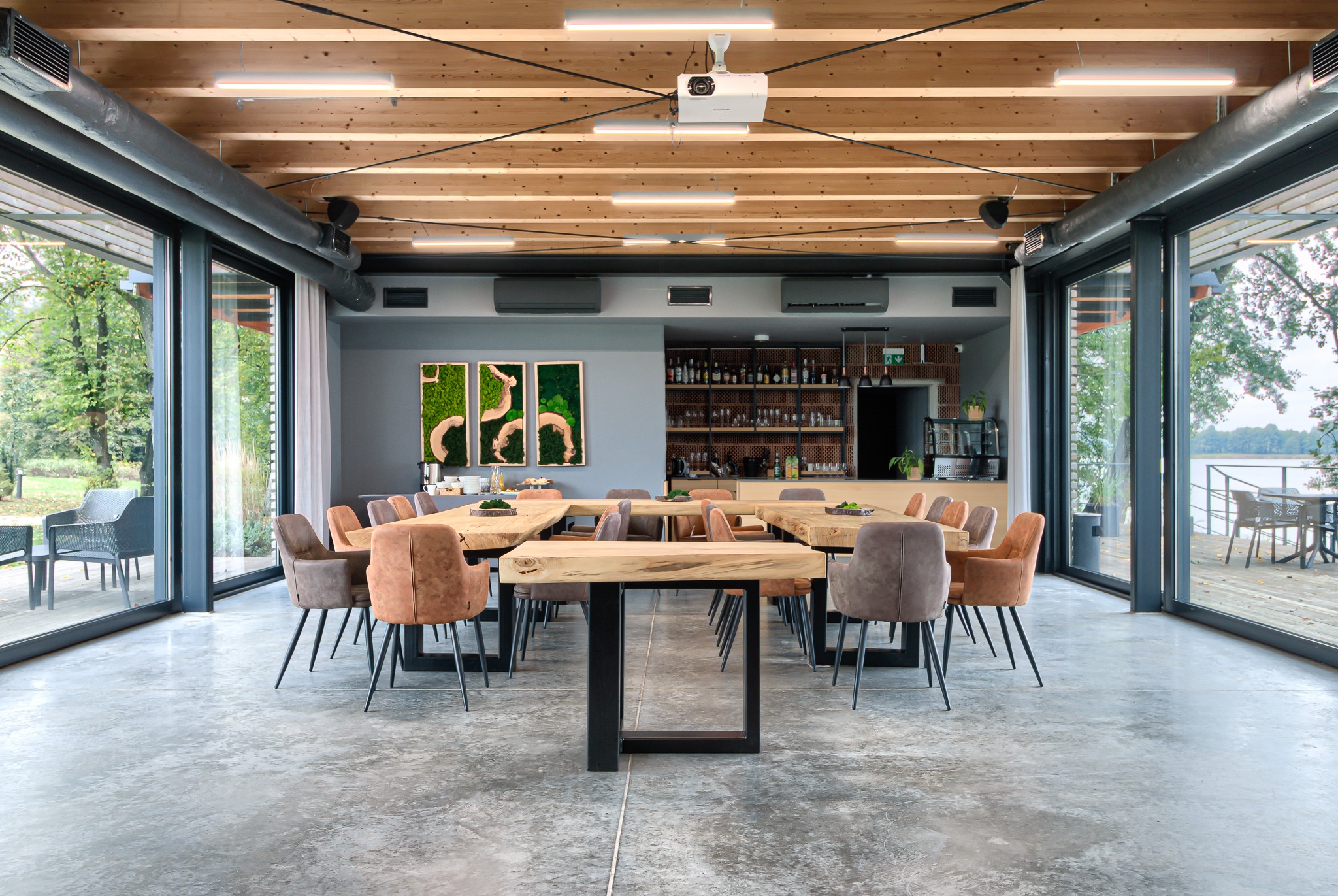 Jasne wnętrze sali konferencyjnej z drewnianym sufitem, projektorem, stołami z surowego drewna i krzesłami w odcieniach brązu, widok na zewnątrz przez panoramiczne okna.