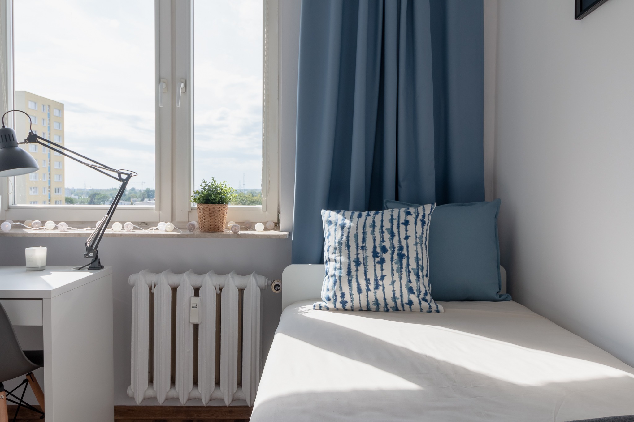 Jasne wnętrze pokoju z łóżkiem, biurkiem, lampką i oknem z widokiem na budynek mieszkalny, dekorowane niebieskimi zasłonami i poduszkami.