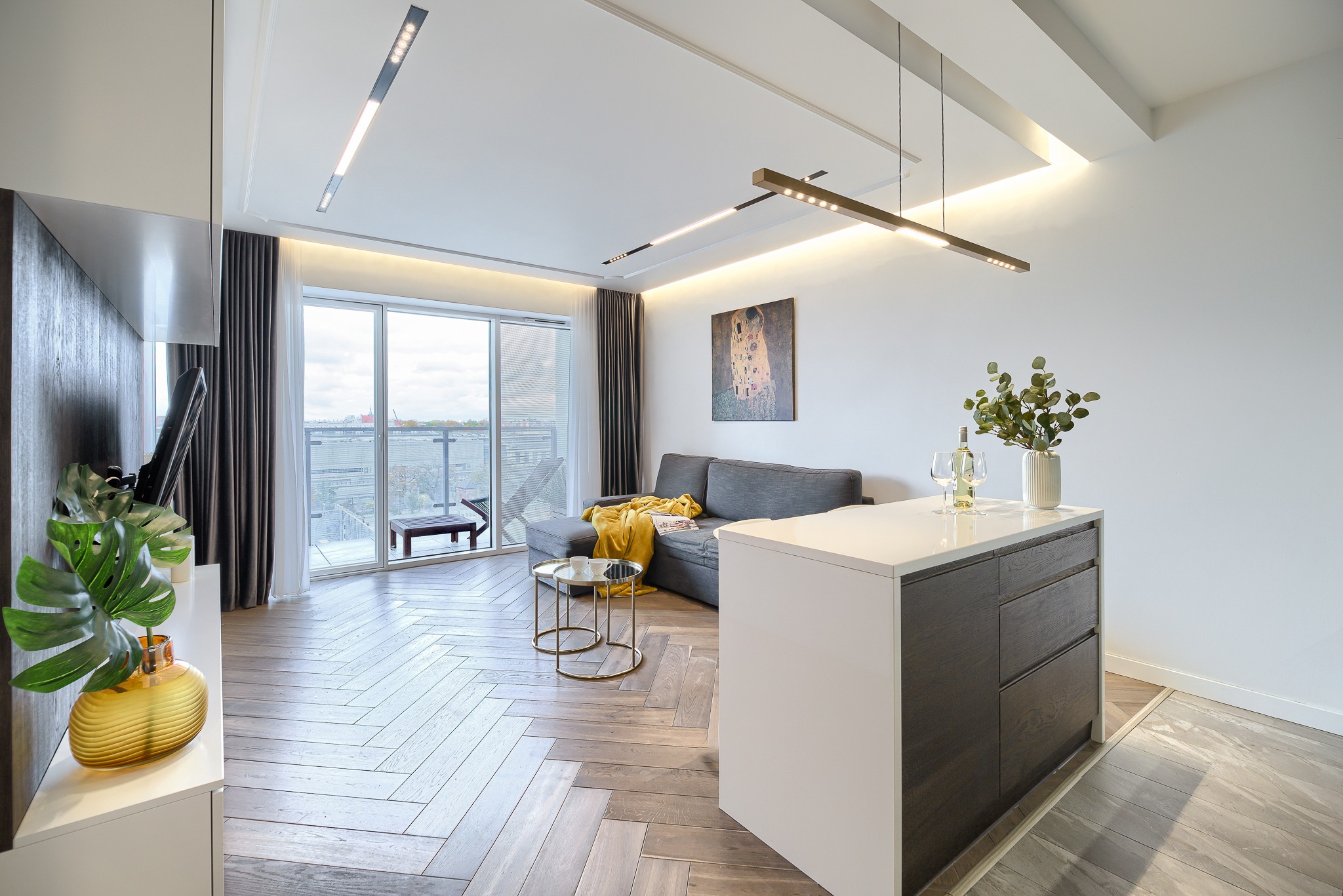Jasne, nowoczesne wnętrze apartamentu z parkietem w jodełkę, szarą sofą z żółtym pledem, wyspą kuchenną i oknem balkonowym z widokiem na miasto.