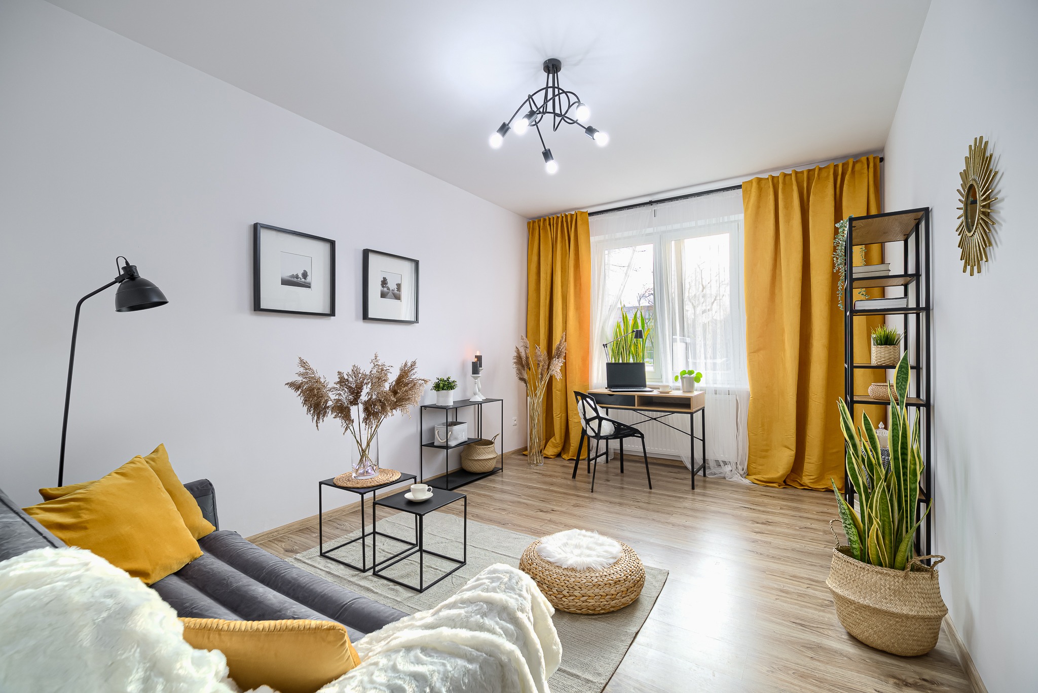 Jasne wnętrze pokoju dziennego z szarą sofą z żółtymi poduszkami, czarną lampą podłogową, stolikami kawowymi, biurkiem przy oknie z żółtymi zasłonami i dekoracyjnymi elementami roślinnymi.