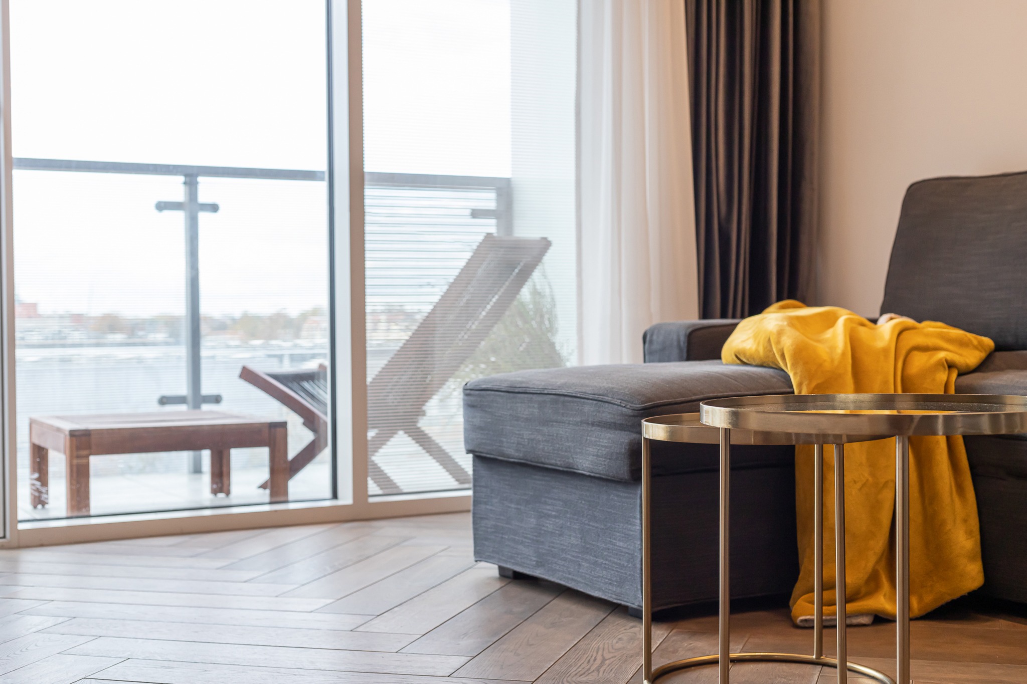Jasne wnętrze z szarą sofą, złotym stolikiem i żółtym pledem, widok na balkon z drewnianymi meblami przez duże okno.