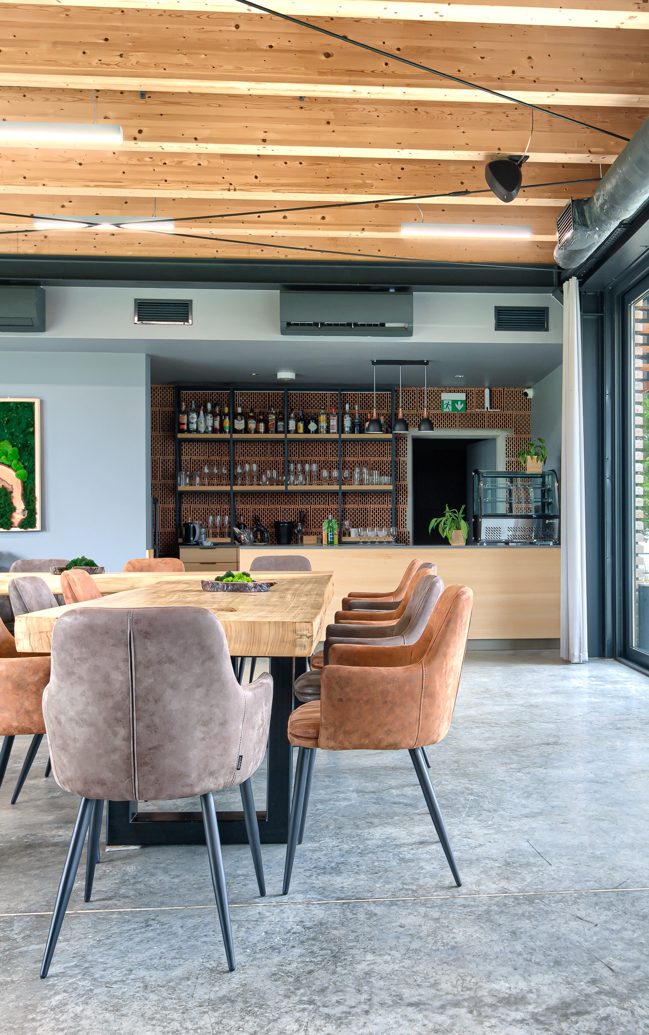 Wnętrze nowoczesnej restauracji lub kawiarni z drewnianym stołem, krzesłami w odcieniach brązu, barem z półkami pełnymi butelek i drewnianym sufitem z widocznymi instalacjami.