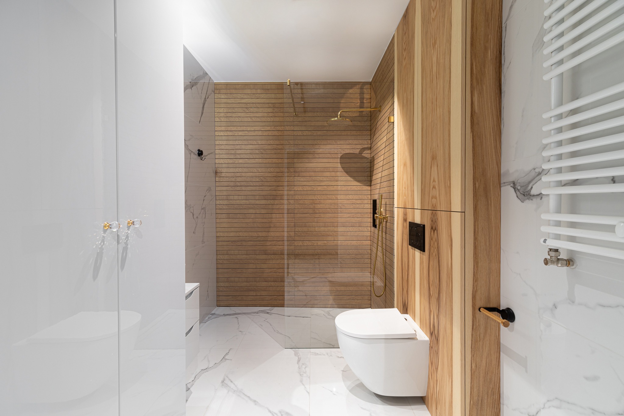 Nowoczesna łazienka z drewnianymi panelami w strefie prysznica, białymi marmurowymi płytkami i złotą armaturą.