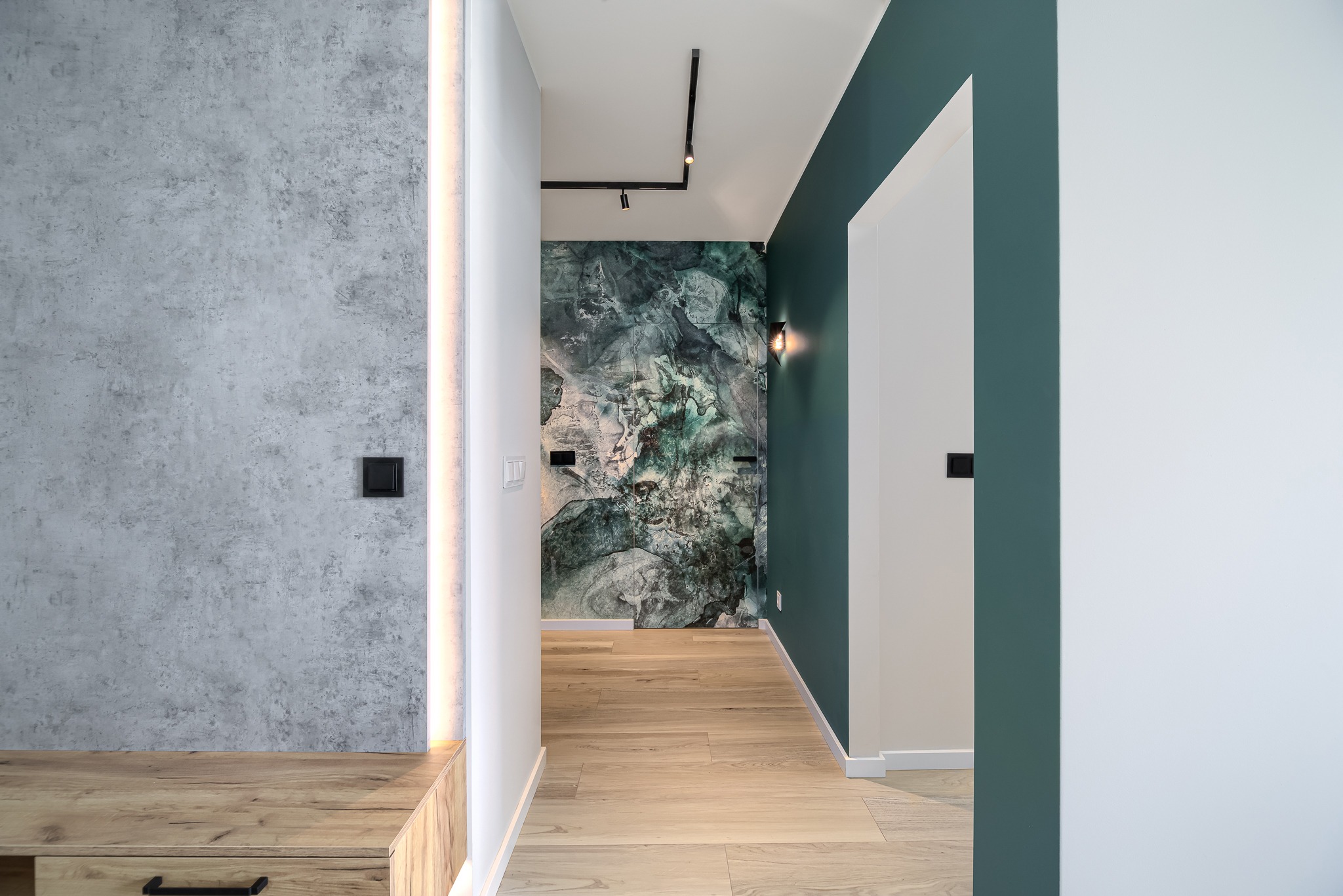Wnętrze korytarza z drewnianą podłogą, betonową ścianą z podświetleniem LED, ścianą z abstrakcyjnym wzorem w odcieniach zieleni i oświetleniem szynowym.