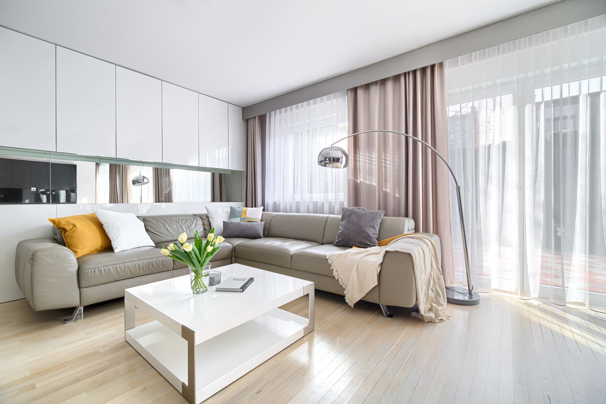 Jasny salon z narożną skórzaną sofą, białym stolikiem kawowym, tulipanami w wazonie, lampą łukową i zasłonami w odcieniach beżu i bieli.