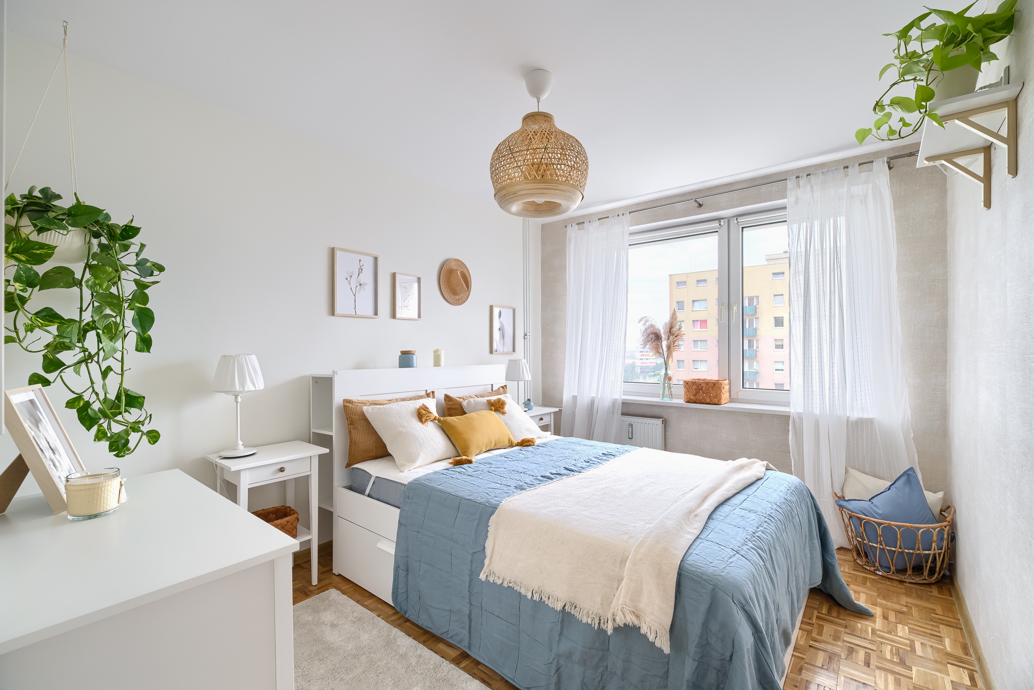 Jasna sypialnia z białymi meblami, łóżkiem z niebieską narzutą i dekoracjami z naturalnych materiałów, takimi jak wiklinowa lampa i kosz.