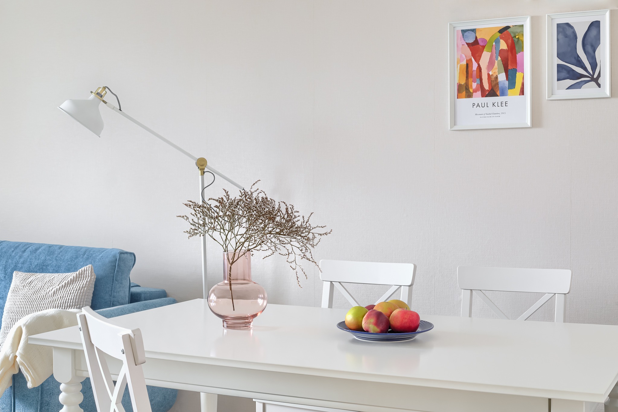 Jasne wnętrze z białym stołem, krzesłami, niebieską kanapą i dekoracjami, wazon z suszem i owoce na talerzu, minimalistyczny wystrój z plakatami na ścianie.