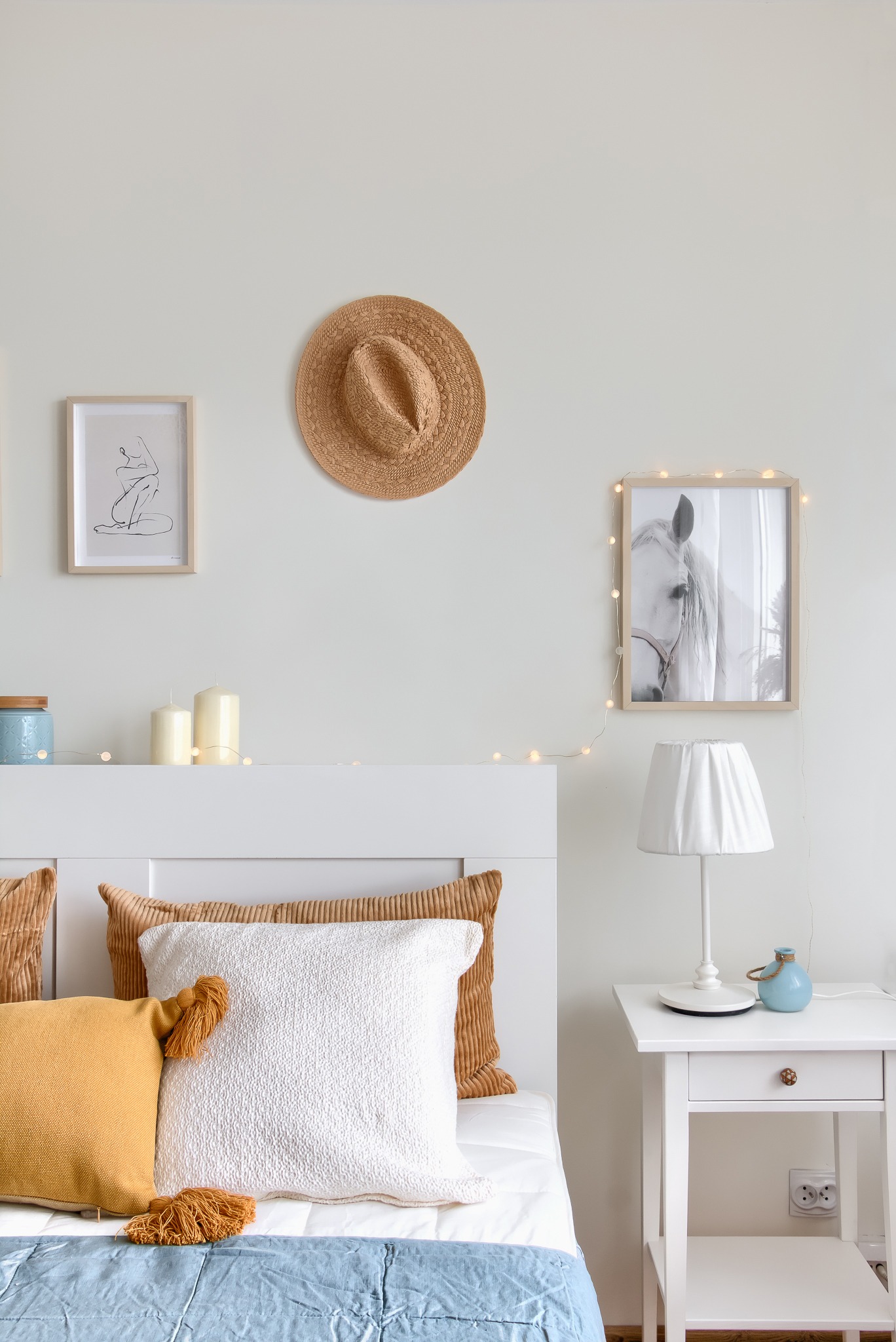 Jasne wnętrze sypialni z białym łóżkiem, dekoracyjnymi poduszkami, lampką nocną na białej szafce, obrazem konia, kapeluszem na ścianie i delikatnymi światełkami.