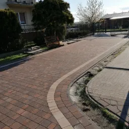 Ułożona z brązowej kostki brukowej ścieżka z jasnymi obrzeżami, prowadząca w kierunku metalowej bramy wjazdowej i domu z balkonem, z widocznymi elementami krajobrazu.