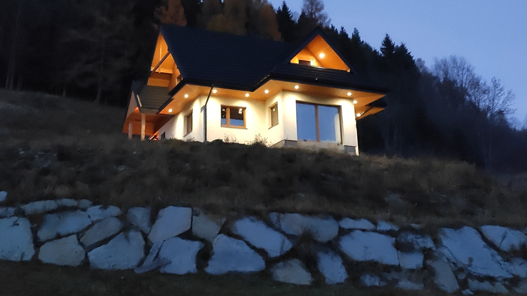 Nowoczesny dom z białymi ścianami i ciemnym dachem, oświetlony od zewnątrz, położony na wzgórzu z kamiennym murem oporowym u podnóża, sfotografowany o zmierzchu.