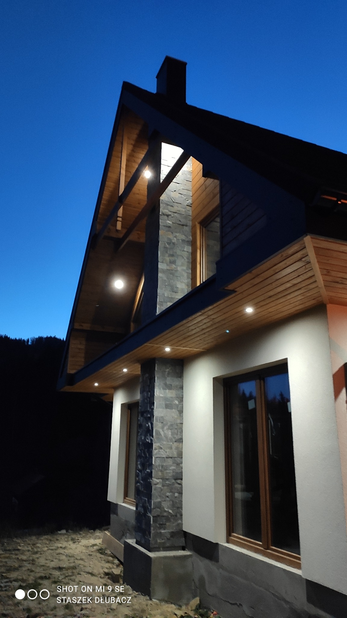 Dół domu szkieletowego z kamienną kolumną, oświetleniem punktowym i drewnianą konstrukcją dachu, sfotografowany o zmierzchu.