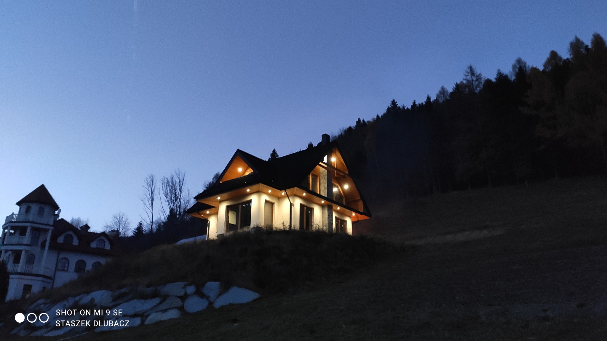 Oświetlony dom w stylu góralskim na wzgórzu, widoczny o zmierzchu z innym budynkiem w tle.